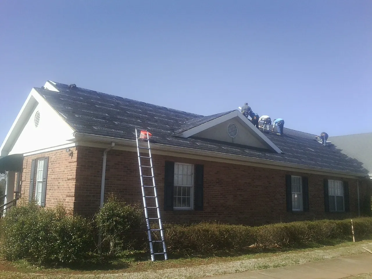 Crew tearing shingles off a brick ranch roof with a ladder set — Metal Roof Repair work by our crew in Riceville